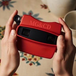 Könnte beinhalten: Ein roter Vintage-Betrachter mit dem Text "MG&CD" wird vor einer floralen Tischdecke gehalten. Der Betrachter hat einen schwarzen Mittelteil und eine kreisförmige Linse. Eine weiße Teekanne und Tasse sind im Hintergrund.