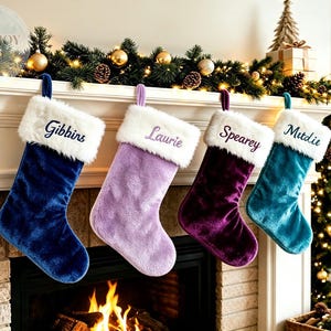 Könnte beinhalten: Vier Plüsch-Weihnachtsstrümpfe in Marineblau, Lavendel, Dunkellila und Blaugrün, jeweils mit einem weißen Kunstfellbündchen. Die Bündchen sind mit den Namen Gibbins, Laurie, Spearey und Mitdie in dunkelblauer oder violetter Schreibschrift bestickt.