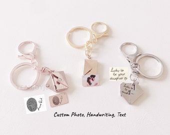 May include: Three keychains with different charms. The first keychain has a gold-colored key ring and a charm that looks like a closed envelope with a small photo inside. The second keychain has a silver-colored key ring and a charm that looks like a folded piece of paper with the text "Lucky to be your daughter" written on it. The third keychain has a silver-colored key ring and a charm that looks like a folded piece of paper with the text "Lucky to be your daughter" written on it. The charms are all attached to the key rings with a small metal loop.