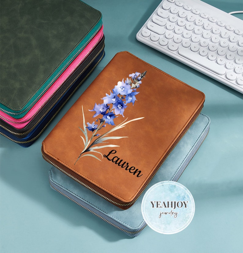 May include: A stack of zippered leather cases in various colors, including green, black, and blue. A brown case with a floral design and the name "Lauren" is in the foreground. A white keyboard is in the background.