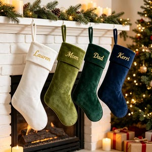 May include: Four velvet Christmas stockings hanging in a row, featuring a white one embroidered with Lauren, an olive green one with Mom, a dark green one with Dad, and a navy blue one with Kerri, each with a matching cuff and hanging loop.