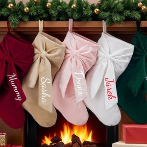 Puede incluir: Cinco calcetines navideños de terciopelo en burdeos, beige, rosa, blanco y verde cuelgan sobre una chimenea. Cada calcetín tiene un lazo grande y un nombre bordado en la parte delantera: Mummy, Sasha, Donna, Jarek y Daddy.