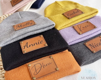 May include: A collection of personalized beanies in various colors, including pink, gray, black, and yellow. Each beanie features a rectangular leather patch with a name engraved on it. The beanies are arranged on a white surface.