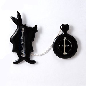 May include: Black enamel pin set featuring a silhouette of the White Rabbit from Alice in Wonderland and a pocket watch. The pins are connected by a silver chain and both feature the signature "Ortiz Sadler" on the back.