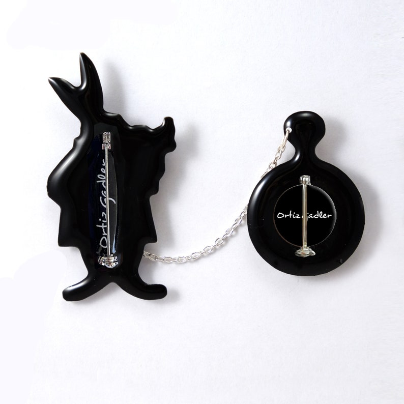 May include: Black enamel pin set featuring a silhouette of the White Rabbit from Alice in Wonderland and a pocket watch. The pins are connected by a silver chain and both feature the signature "Ortiz Sadler" on the back.