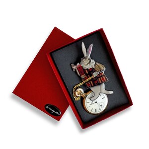 May include: A white rabbit with a pocket watch and a red and black vest, holding a book with playing cards. The rabbit is standing on a pocket watch with Roman numerals. The image is in a red box with a white label that says "Orla Gilder".
