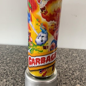 Puede incluir: Un vaso de acero inoxidable con un diseño colorido que presenta personajes de dibujos animados y las palabras "Garbage Pail Kids" y "GPK". El vaso está sobre una base giratoria.