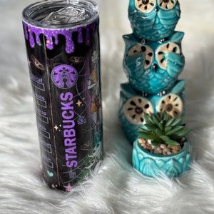 May include: A black Starbucks tumbler with a purple glitter rim and a Halloween-themed design. The tumbler is next to a stack of three teal owl figurines and a small teal pot with a succulent.