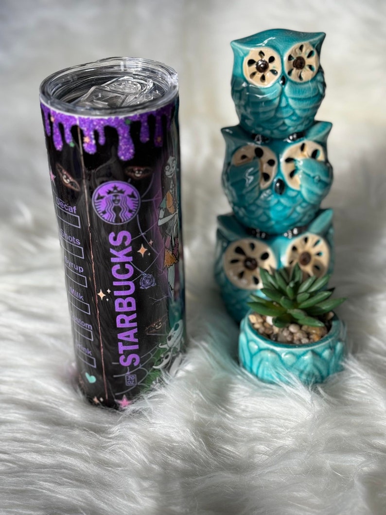 May include: A black Starbucks tumbler with a purple glitter rim and a Halloween-themed design. The tumbler is next to a stack of three teal owl figurines and a small teal pot with a succulent.