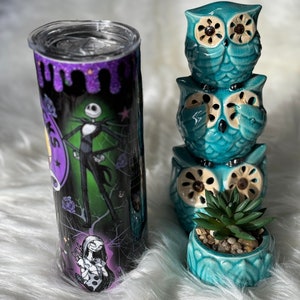 May include: A stainless steel tumbler featuring Jack Skellington and Sally from The Nightmare Before Christmas, with a black, purple, and green design. Beside it are three stacked turquoise owl figurines and a small succulent in a matching pot.