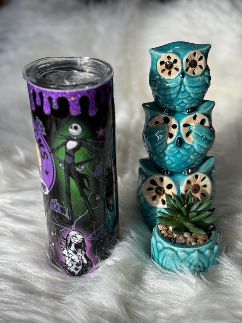 May include: A stainless steel tumbler featuring Jack Skellington and Sally from The Nightmare Before Christmas, with a black, purple, and green design. Beside it are three stacked turquoise owl figurines and a small succulent in a matching pot.