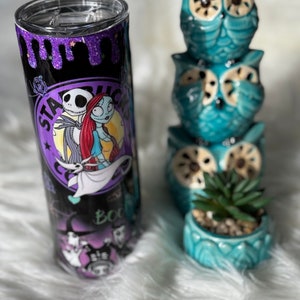 May include: A stainless steel tumbler featuring a design with Jack Skellington and Sally from The Nightmare Before Christmas. The tumbler has a black background with purple glitter accents and the Starbucks logo. Beside the tumbler are three stacked teal owl figurines and a small teal pot with a succulent.