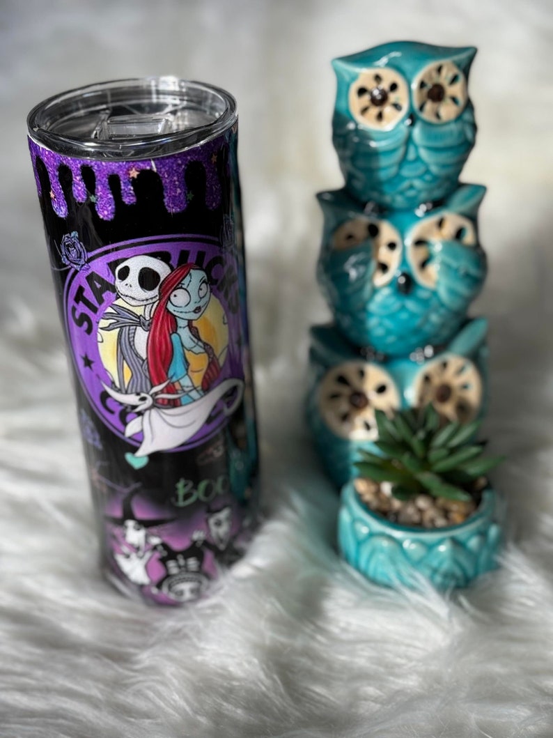 May include: A stainless steel tumbler featuring a design with Jack Skellington and Sally from The Nightmare Before Christmas. The tumbler has a black background with purple glitter accents and the Starbucks logo. Beside the tumbler are three stacked teal owl figurines and a small teal pot with a succulent.