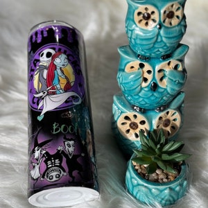 May include: A stainless steel tumbler featuring characters from The Nightmare Before Christmas, with a black and purple background. Next to it are three stacked teal ceramic owl figurines and a small teal planter with a succulent.