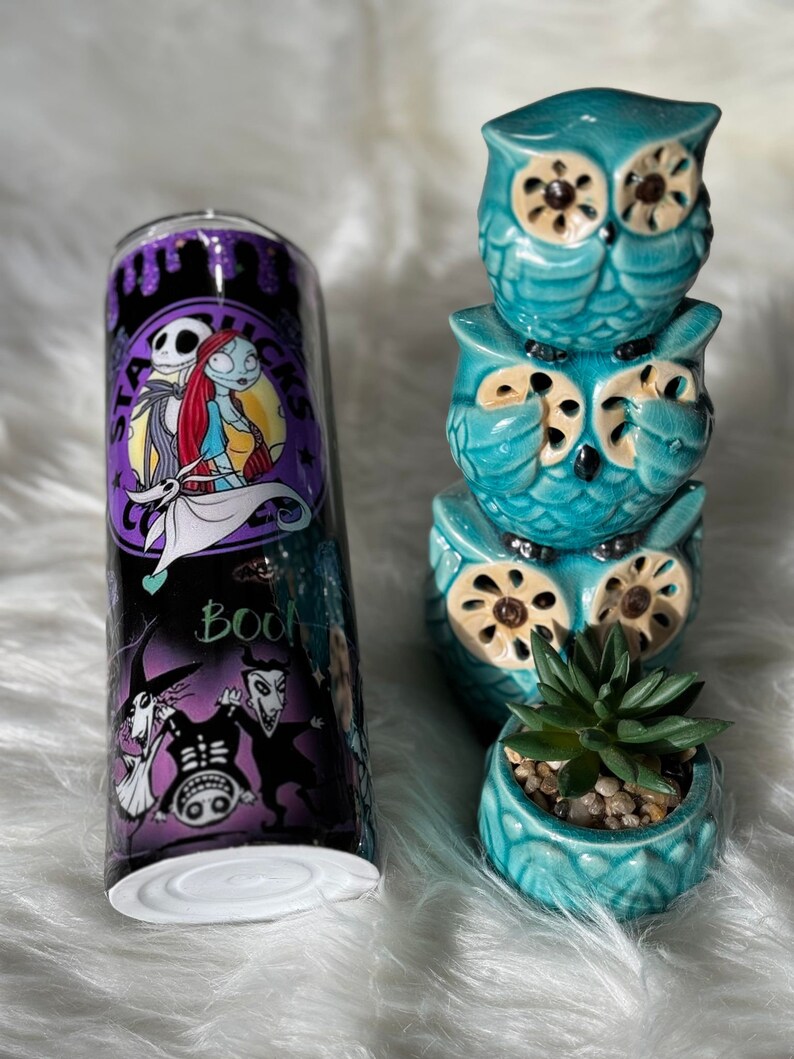May include: A stainless steel tumbler featuring characters from The Nightmare Before Christmas, with a black and purple background. Next to it are three stacked teal ceramic owl figurines and a small teal planter with a succulent.