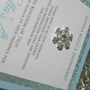 May include: A white invitation with a blue border and silver glitter. The invitation is for a birthday party and says "Wanna build a snowman?"  The invitation also says "Please join us as our princess turns another year older! The gates of Addington Lane are opening for celebration day. The Kingdom of Troy requests the honor of your presence for Ella's 7th"