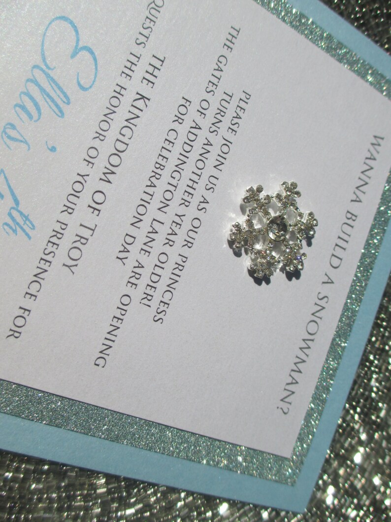 May include: A white invitation with a blue border and silver glitter. The invitation is for a birthday party and says "Wanna build a snowman?"  The invitation also says "Please join us as our princess turns another year older! The gates of Addington Lane are opening for celebration day. The Kingdom of Troy requests the honor of your presence for Ella's 7th"