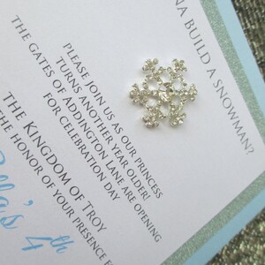 May include: A light blue invitation with silver glitter trim. The invitation features a silver snowflake with a clear rhinestone center. The text on the invitation reads "Wanna build a snowman?" and includes details about a birthday party for a princess turning four years old.