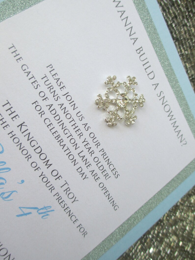 May include: A light blue invitation with silver glitter trim. The invitation features a silver snowflake with a clear rhinestone center. The text on the invitation reads "Wanna build a snowman?" and includes details about a birthday party for a princess turning four years old.
