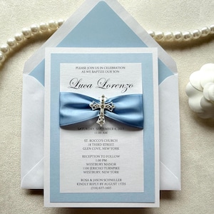 May include: A baptism invitation card for Luca Lorenzo featuring a light blue satin ribbon and a silver cross charm with clear rhinestones. The white card has black text detailing the event, date, time, and locations in New York, set against a matching light blue envelope.