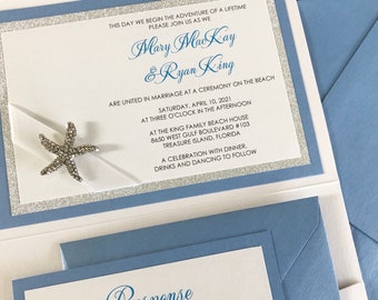 May include: A light blue wedding invitation set featuring a white card with silver glitter borders and a rhinestone starfish embellishment. The text reads: This day we begin the adventure of a lifetime please join us as we Mary MacKay & Ryan King.