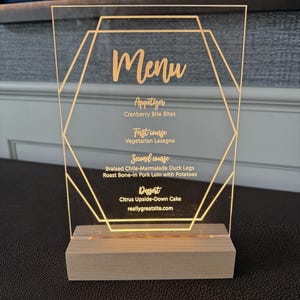 Op de afbeelding: Een helder acrylmenu met een gouden geometrisch ontwerp en een houten voet. Het menu bevat de gangen: Voorgerecht, Eerste gang, Tweede gang en Dessert. Het menu bevat de tekst "Menu" en de website reallygreatsite.com.