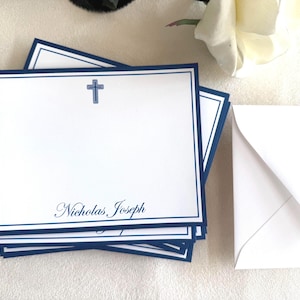 Pode incluir: Uma pilha de cartões de papelaria brancos com uma cruz azul e o nome "Nicholas Joseph" impresso em escrita elegante. Cada cartão tem uma borda azul marinho. Um envelope branco está ao lado dos cartões.