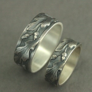 May include: Two silver wedding bands with an oak leaf and acorn design. The bands are textured and have a dark oxidized finish.