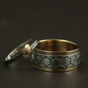 May include: Two wedding rings. One ring is silver with a gold bee accent. The other ring is silver with a honeycomb pattern and a gold interior. The rings are set against a dark background.