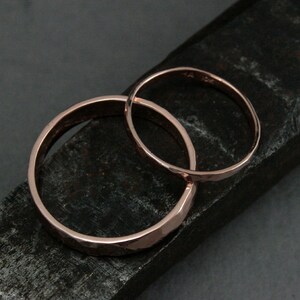 May include: Two hammered rose gold wedding bands, one wider than the other, are stacked on a dark gray surface.