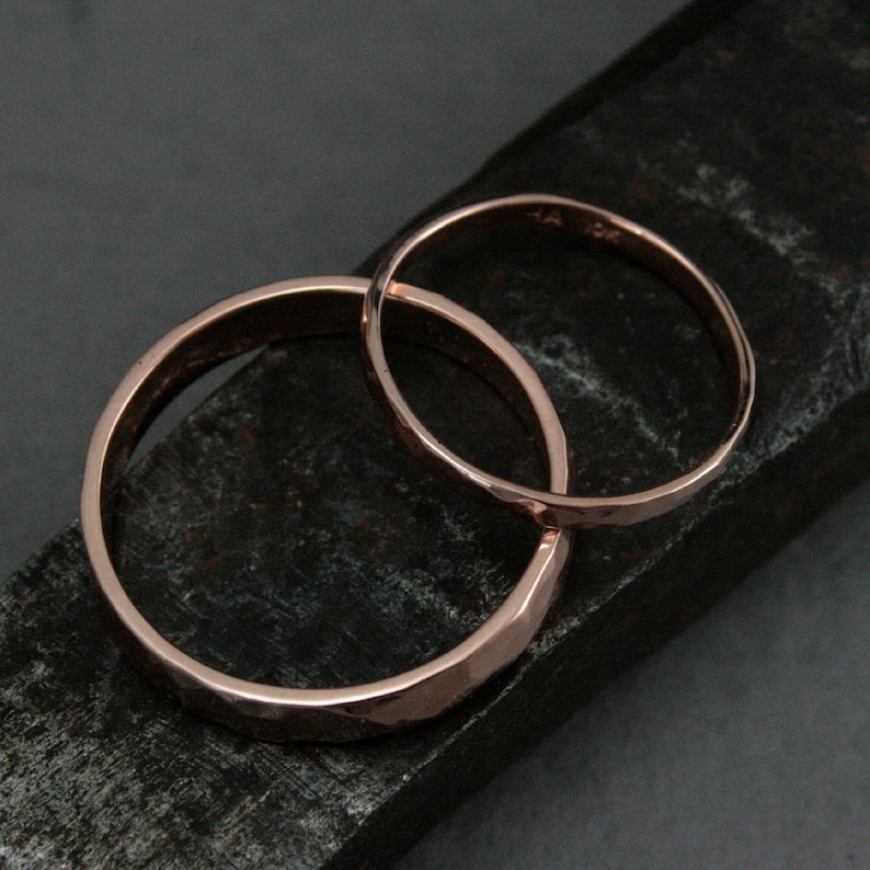 May include: Two hammered rose gold wedding bands, one wider than the other, are stacked on a dark gray surface.
