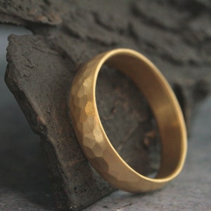 May include: A gold band ring with a hammered texture. The ring is sitting on a piece of rough, dark brown wood.