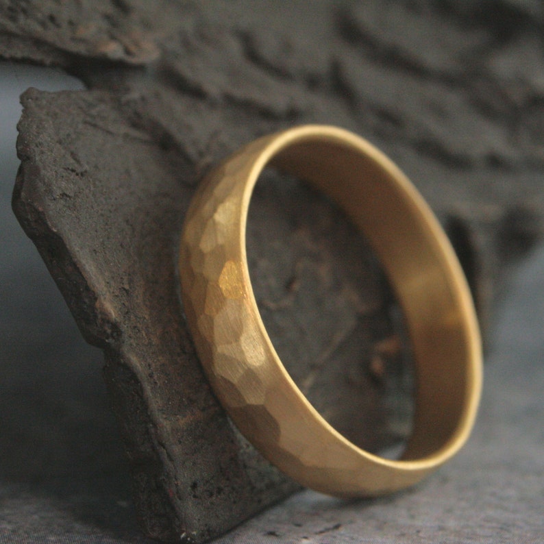 May include: A gold band ring with a hammered texture. The ring is sitting on a piece of rough, dark brown wood.