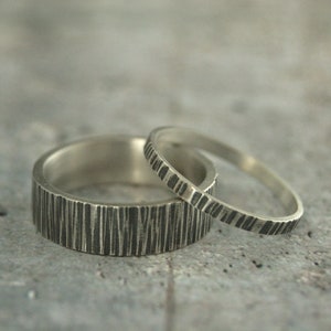 May include: Two silver rings with a textured, bark-like pattern. The larger ring has a wider band, while the smaller ring is more slender. Both rings are set against a neutral gray background.