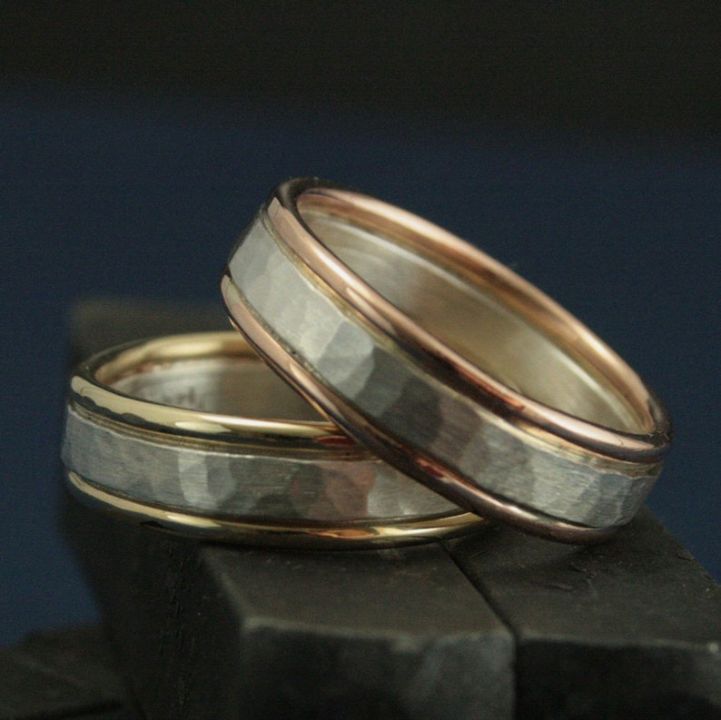 May include: Two wedding bands with a hammered finish. The top band is silver with a rose gold inlay. The bottom band is silver with a yellow gold inlay.
