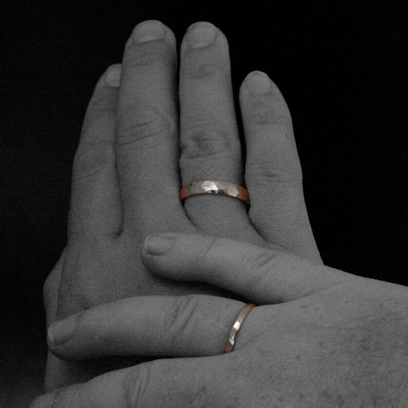 May include: Two hands with wedding bands on their ring fingers. The bands are both gold and have a hammered texture.