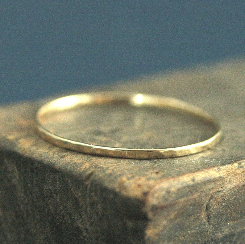 May include: A thin, circular gold ring with a smooth, polished surface. The band features a simple, continuous design with a reflective metallic finish that catches the light, creating a subtle glow along the rounded edge of the jewelry piece.