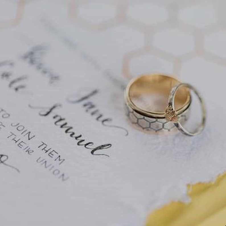 May include: Two wedding rings on a white invitation with handwritten text. One ring is gold with a honeycomb pattern, and the other is a thin, silver band. The invitation has a torn edge and a yellow border.