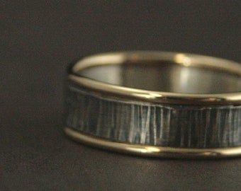 May include: A close-up of a ring with a textured, silver band and gold edges. The ring's band has a vertical, linear pattern, and the gold edges provide a contrasting, polished finish. The ring is set against a dark background.