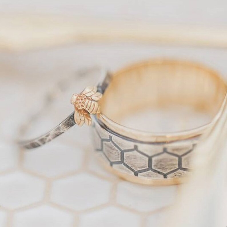 May include: Two rings, one gold and one silver, are displayed. The gold ring has a honeycomb pattern and a gold interior. The silver ring features a small bee design. The rings are likely wedding bands or fashion accessories.