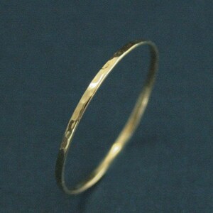 May include: A thin, circular gold ring featuring a hammered, textured surface that creates small, reflective indentations along the band. The metal has a polished, metallic finish that catches the light, highlighting the uneven, hand-crafted appearance.
