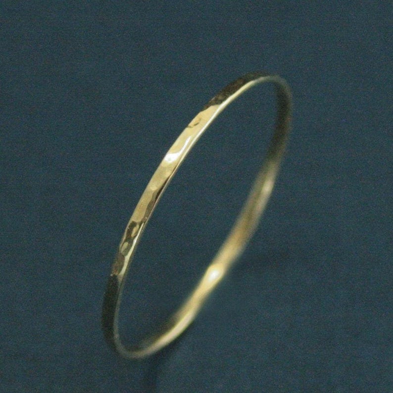 May include: A thin, circular gold ring featuring a hammered, textured surface that creates small, reflective indentations along the band. The metal has a polished, metallic finish that catches the light, highlighting the uneven, hand-crafted appearance.