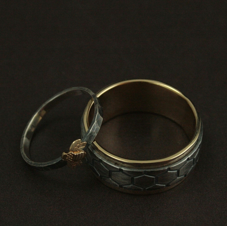 May include: Two rings, one silver and one gold, are displayed against a dark background. The silver ring has a textured surface and a small gold bee charm. The larger ring has a honeycomb pattern and a gold interior.