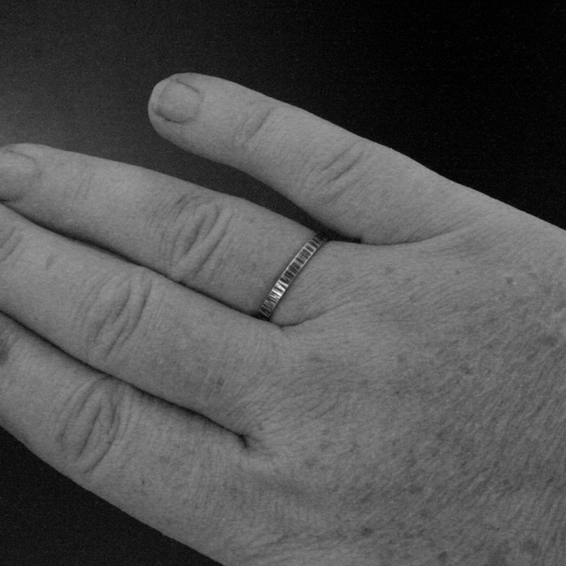 May include: A silver band ring with a textured surface. The ring is worn on a finger, with the hand in the foreground. The ring's design features a series of small, rectangular indentations around the band.