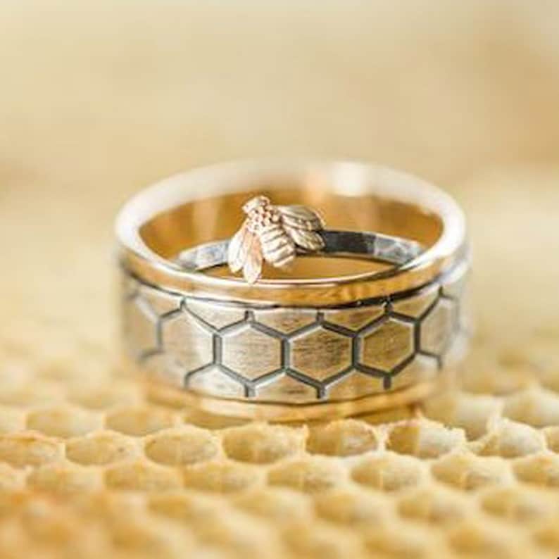 May include: Two rings, one with a gold band and a silver band with a honeycomb pattern, and another with a gold bee on top. The rings are set on a honeycomb surface. The rings are likely wedding or engagement rings.