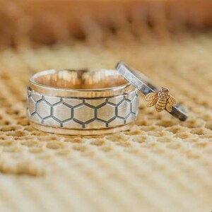 May include: Two rings, one with a honeycomb pattern and the other with a small bee. The larger ring has a gold interior and a silver exterior with a honeycomb design. The smaller ring has a silver band with a gold bee.