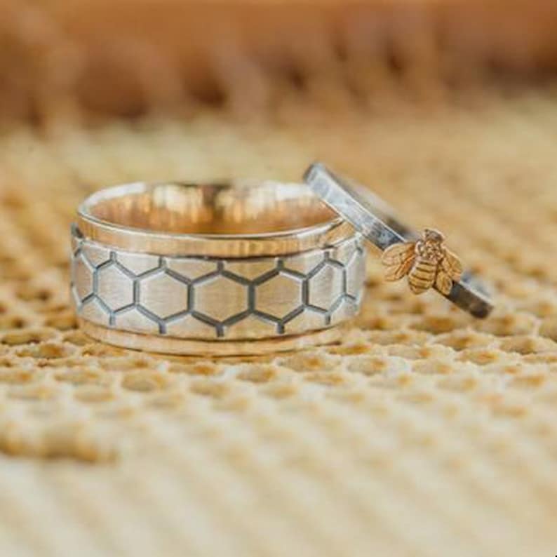 May include: Two rings, one with a honeycomb pattern and the other with a small bee. The larger ring has a gold interior and a silver exterior with a honeycomb design. The smaller ring has a silver band with a gold bee.
