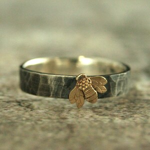 May include: A silver band ring with a hammered finish and a gold bee charm.
