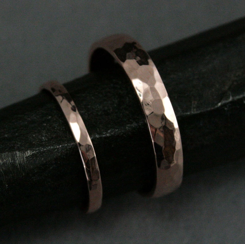 May include: Two hammered rose gold wedding bands, one wider than the other, on a black surface.