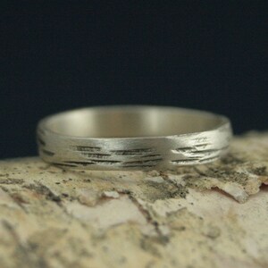 May include: A silver band ring with a textured surface that resembles the bark of a tree.